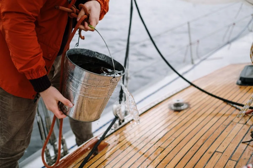 Crew member rinsing a teak yacht deck with fresh water during a washdown